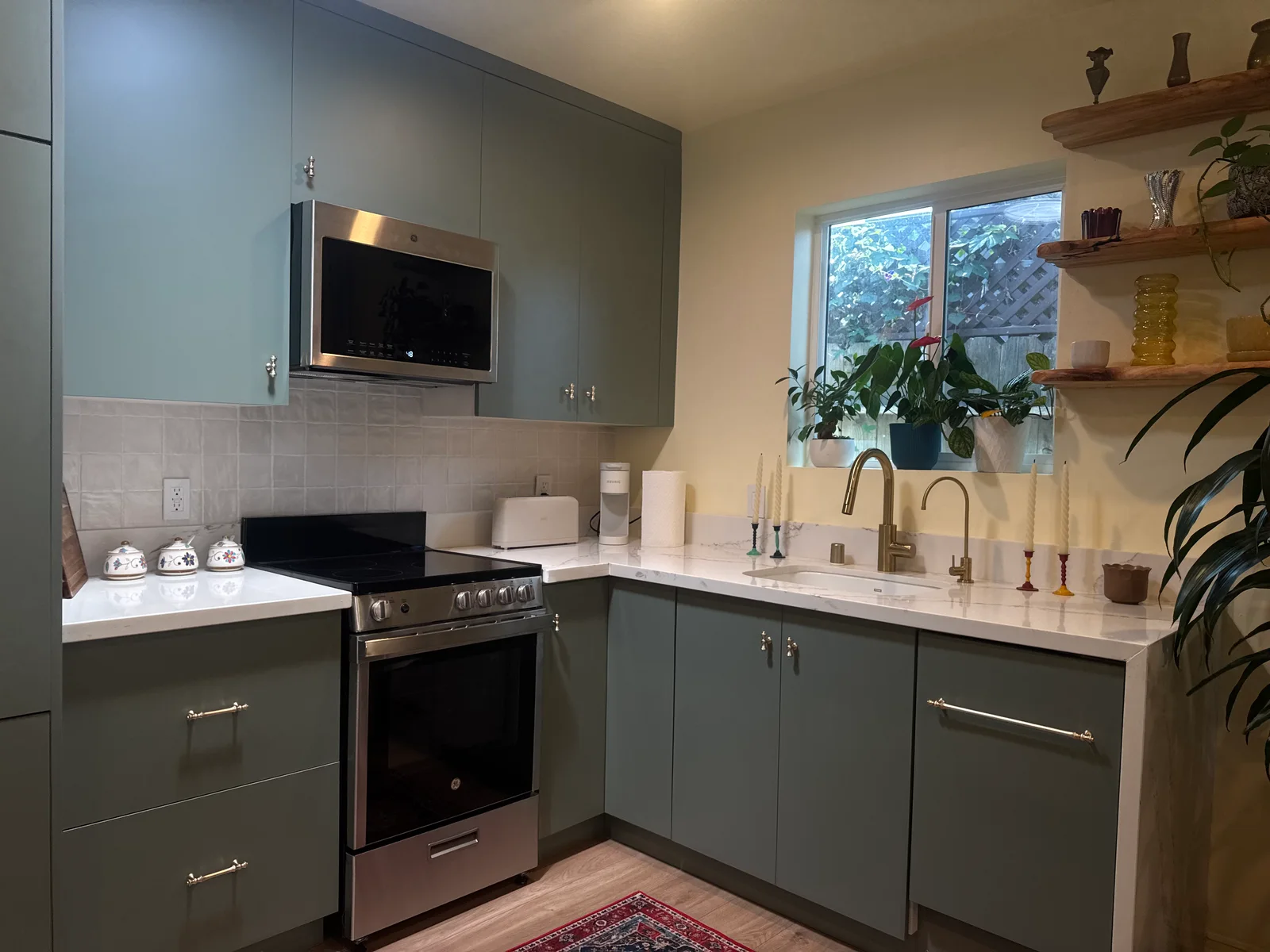 A kitchen with green cabinets, open wooden shelves, and plants