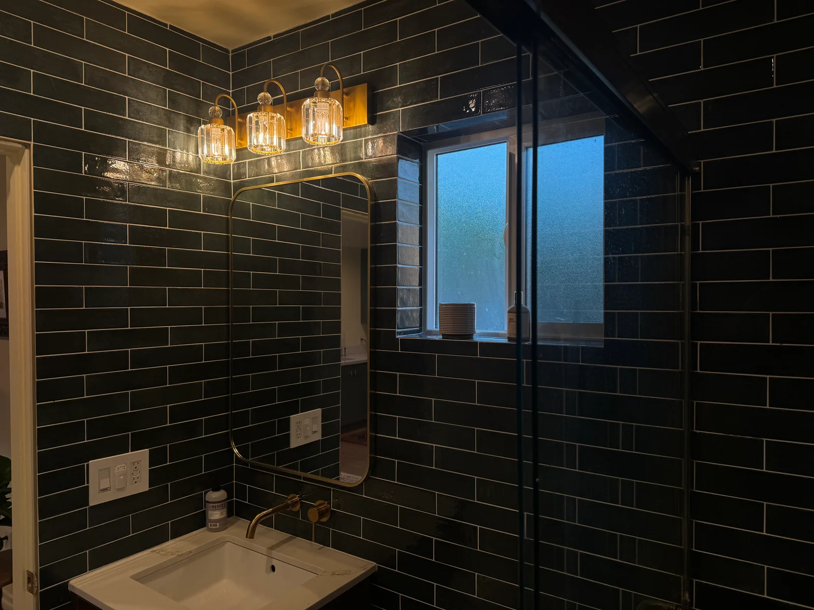 A bathroom with dark tiles, a mirror, and warm pendant lights